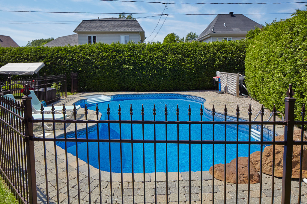 A backyard swimming pool with metal fence on the foreground and hedges on the background