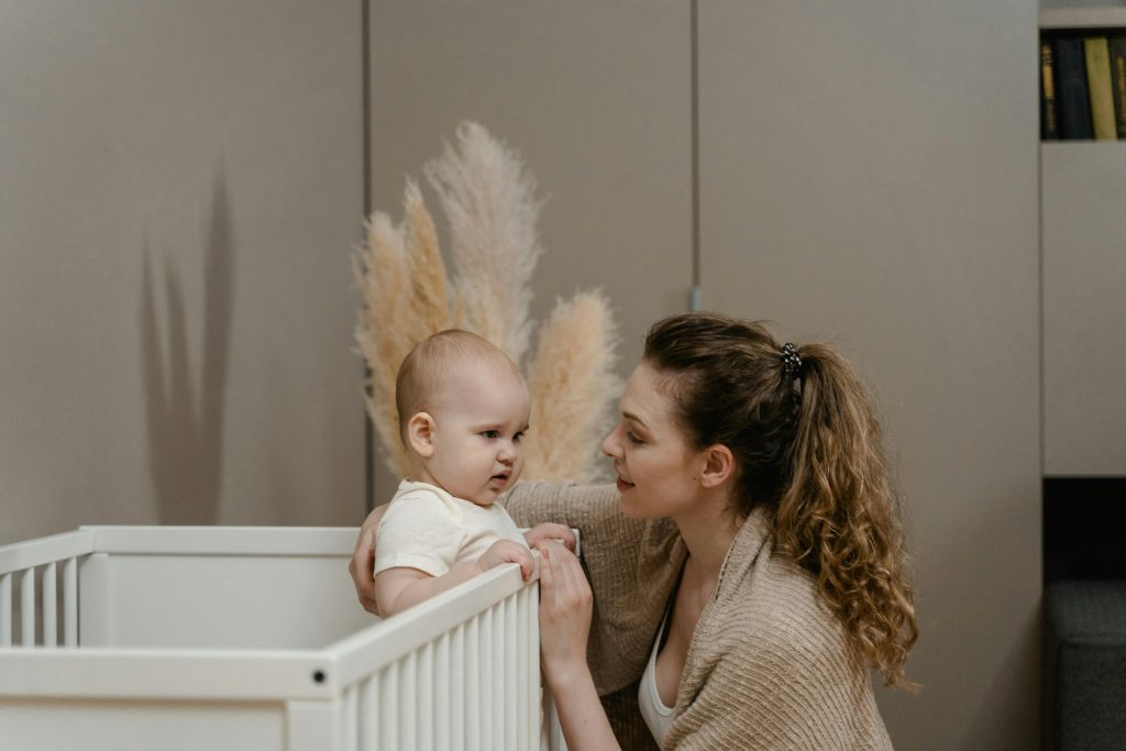 frowning baby standing in a crib with his mother sitting beside him